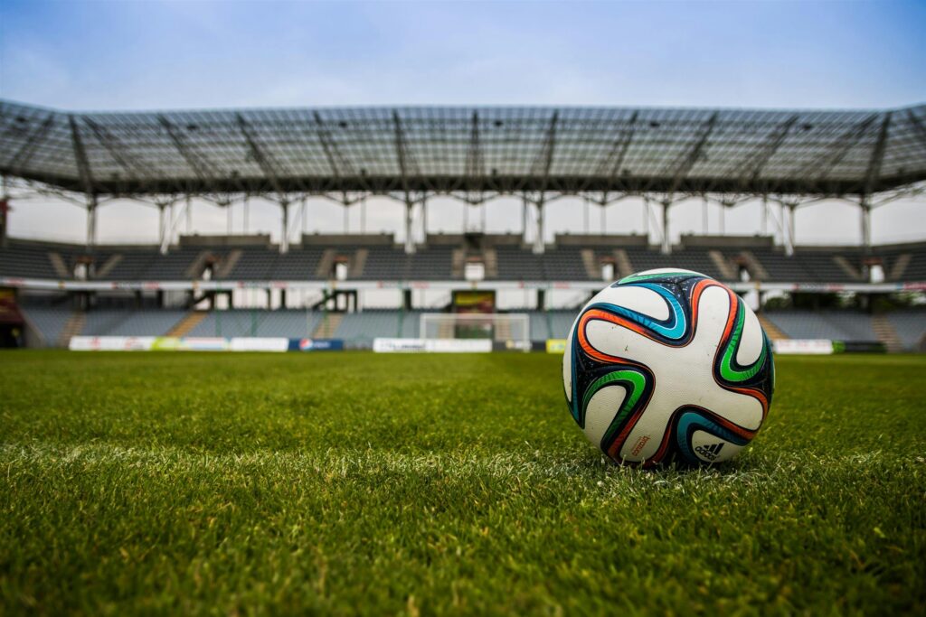 the-ball-stadion-football-the-pitch-46798-46798 Close-up of a soccer ball on a lush grass field with an empty stadium in the background.